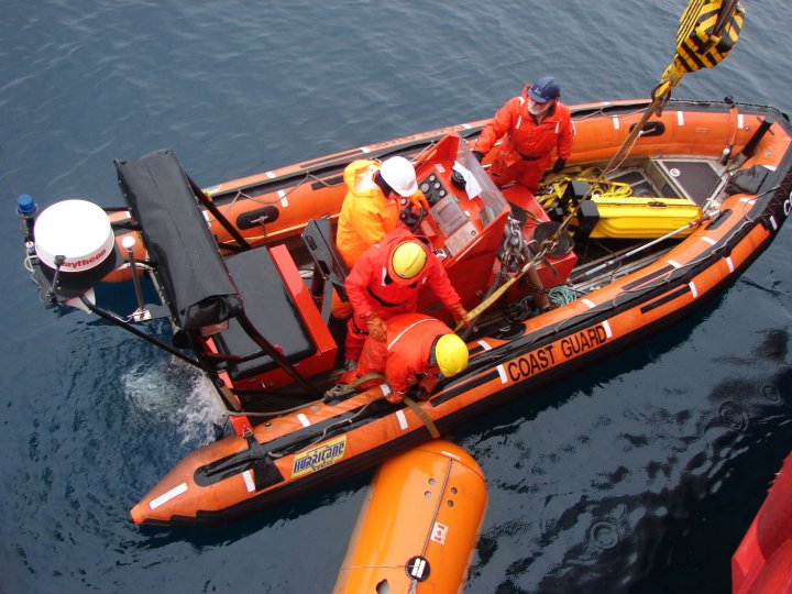 An orange inflatable boat, seen from above. Several figures in hardhats and orange jumpsuits are standing inside.